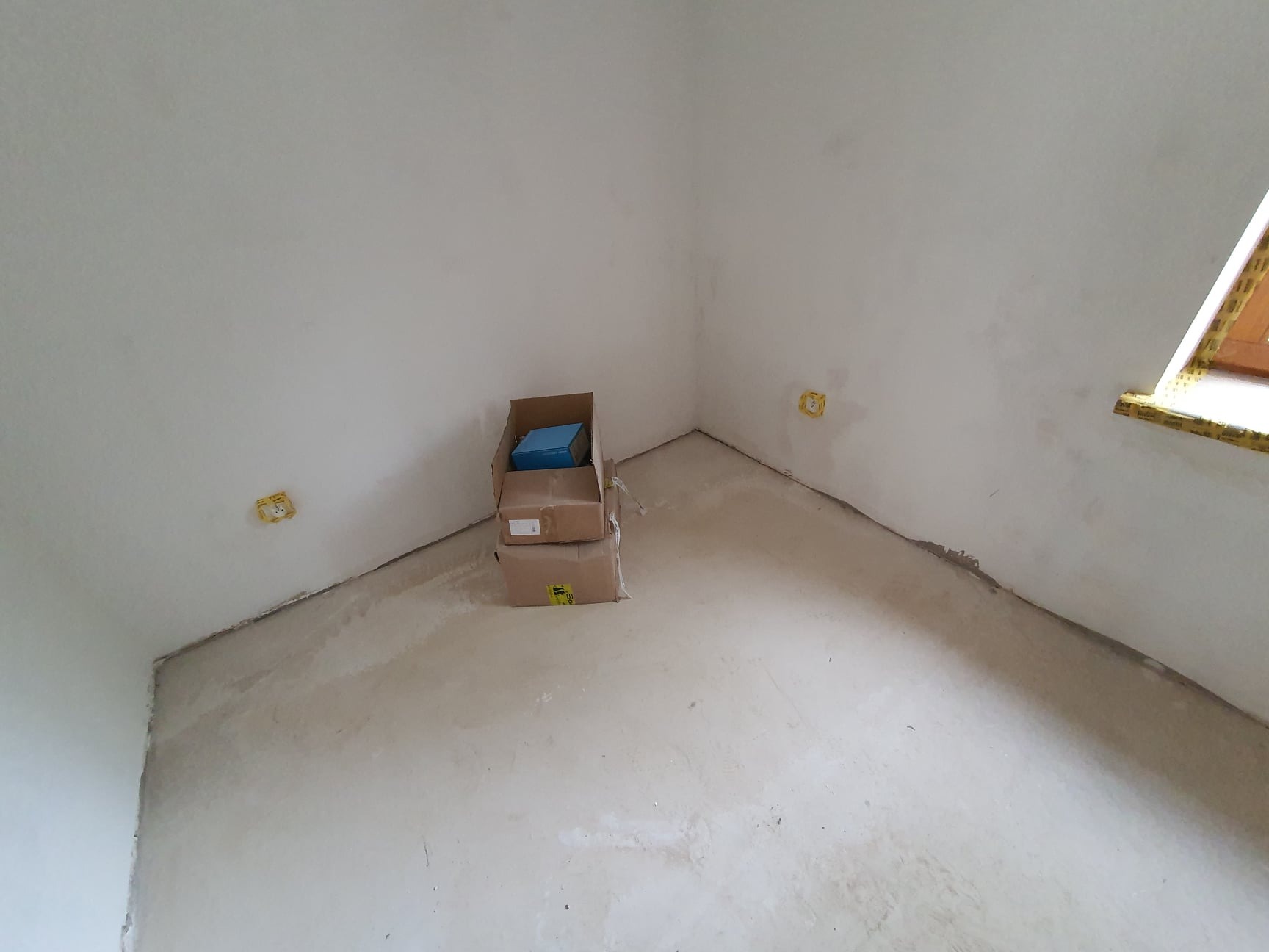 Przygotowanie do prac wykończeniowych w Elblągu: surowe ściany i podłoga po szpachlowaniu, widoczne puszki elektryczne i okno zabezpieczone taśmą, kartony ze sprzętem w rogu pomieszczenia.