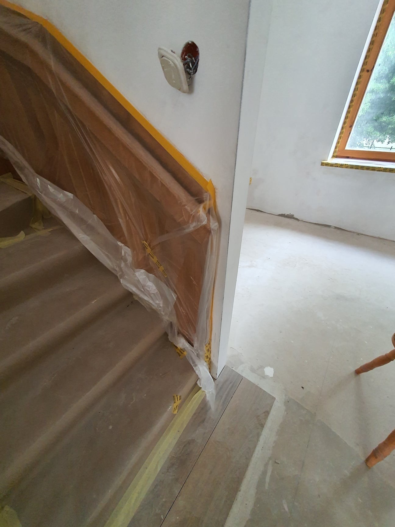 Schody zabezpieczone folią i taśmą malarską podczas prac wykończeniowych, widoczne gniazdko elektryczne i okno w tle.