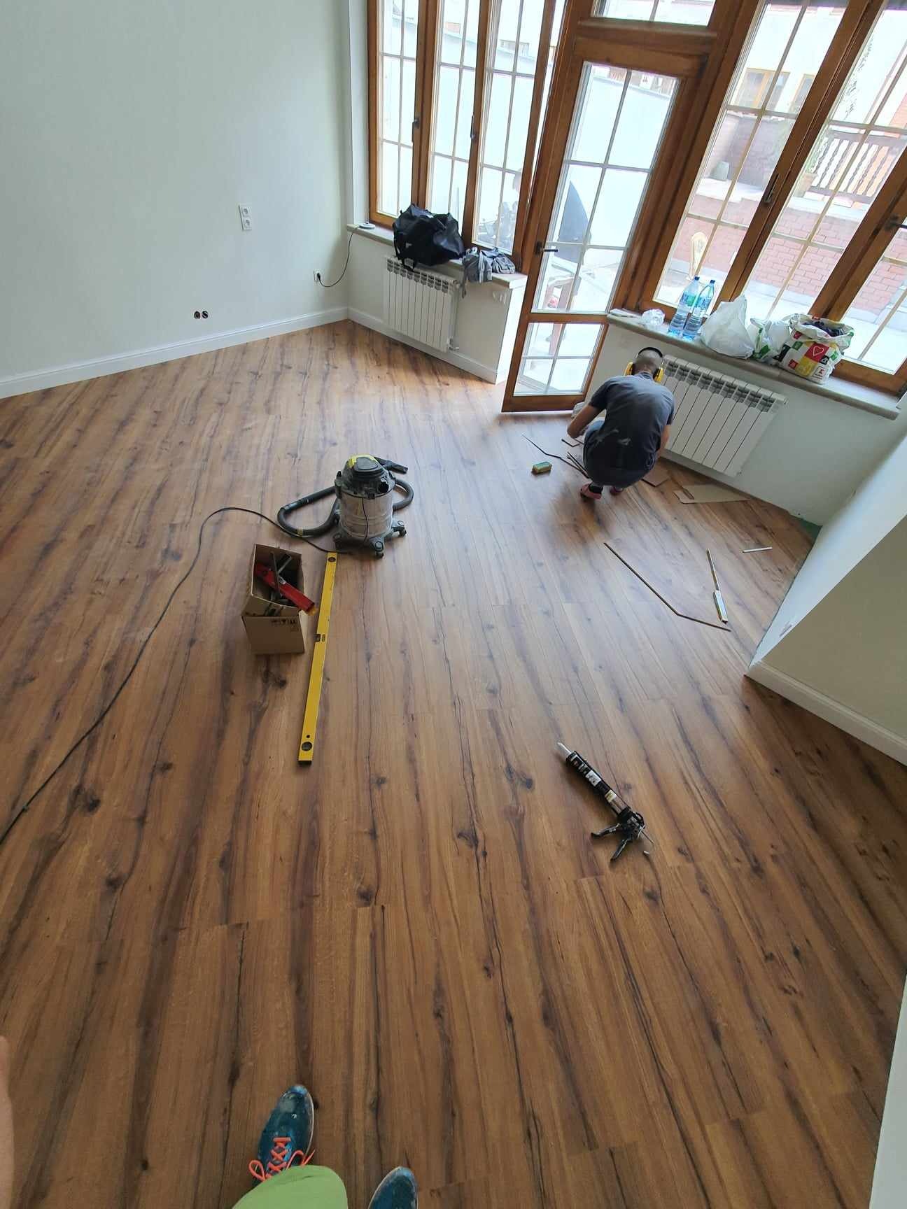 Montaż paneli podłogowych w pokoju z oknami, widoczny pracownik w trakcie pracy, poziomica, odkurzacz i pistolet do kleju.