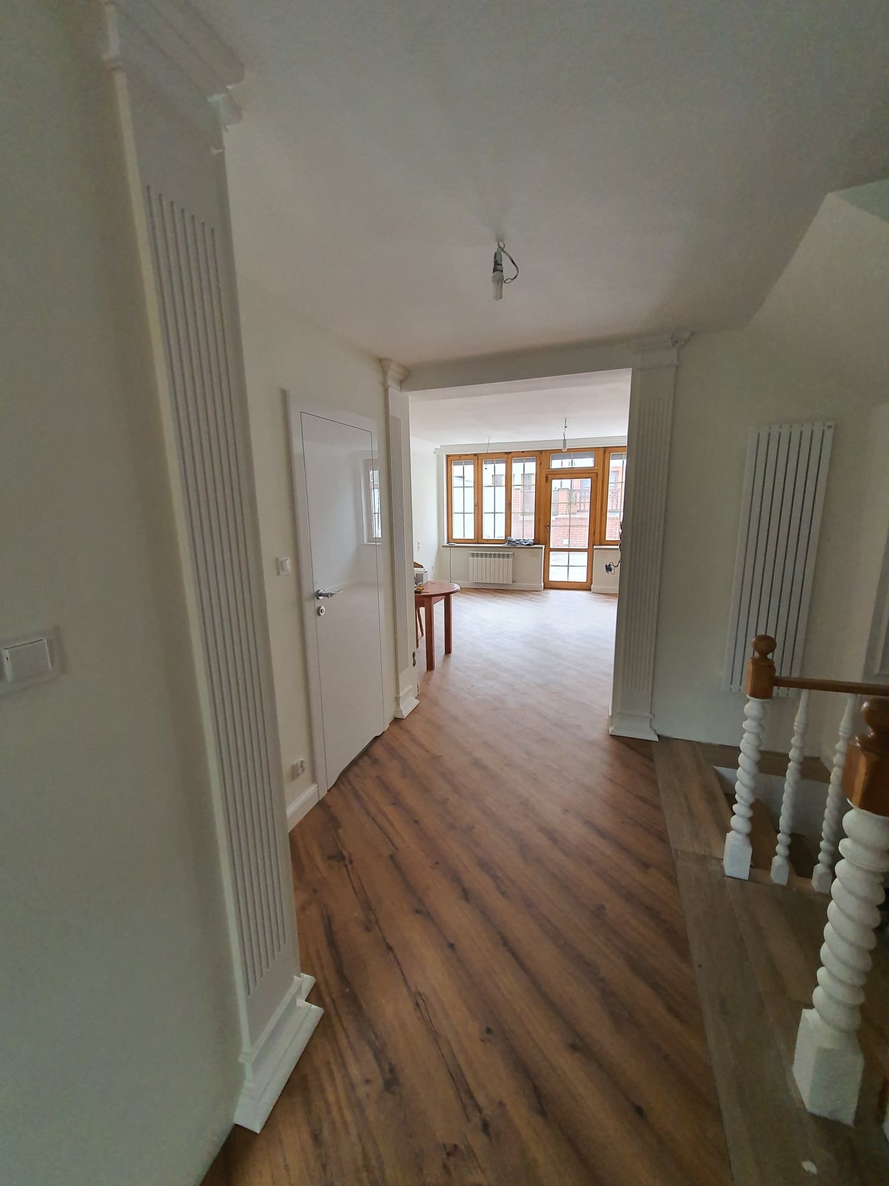 Korytarz z drewnianą podłogą ułożoną w jodełkę, białe ściany z dekoracyjnymi kolumnami i listwami przypodłogowymi, widok na jasny pokój z drewnianymi oknami.