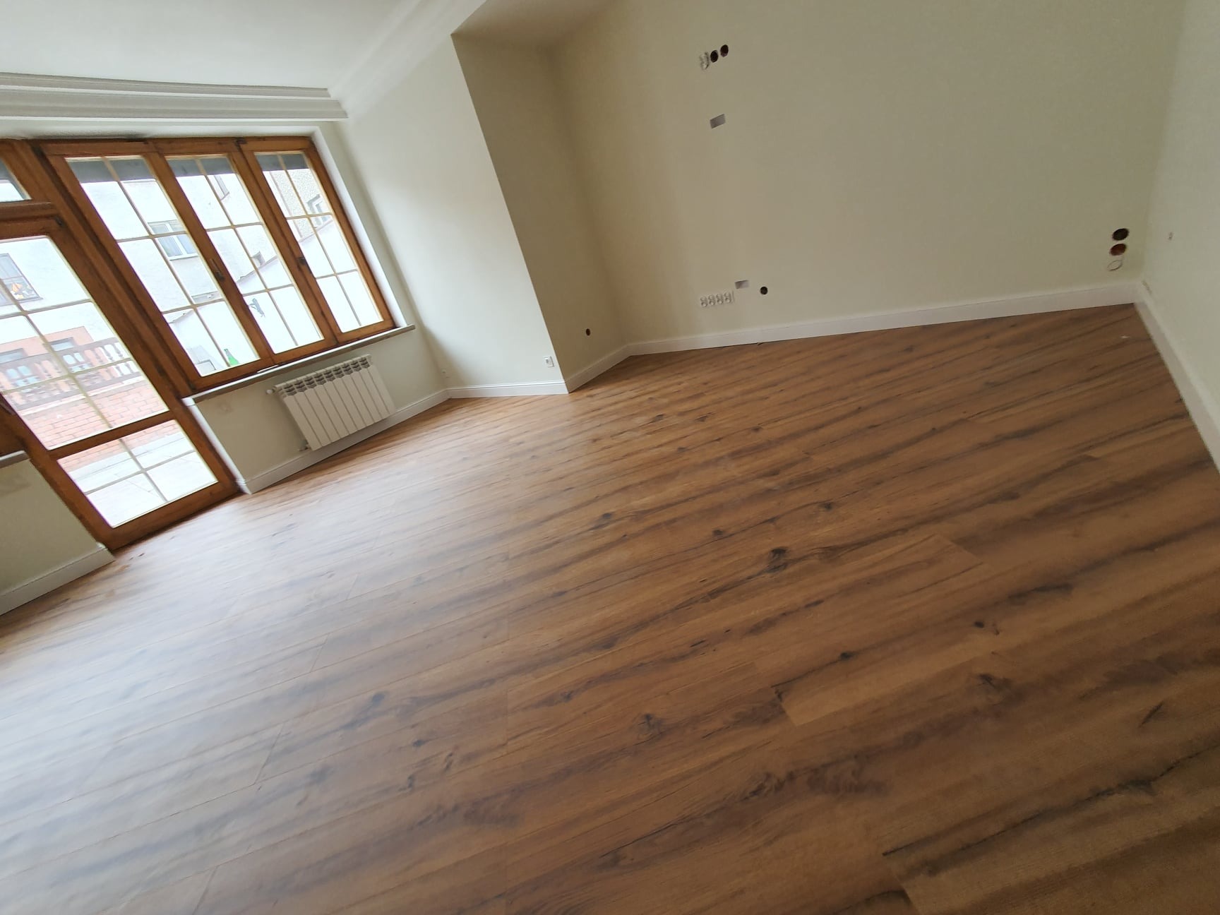 Wykończone wnętrze pokoju z nową podłogą z paneli w odcieniach brązu, jasnymi ścianami z widocznymi gniazdkami i wyłącznikami oraz drewnianą stolarką okienną.