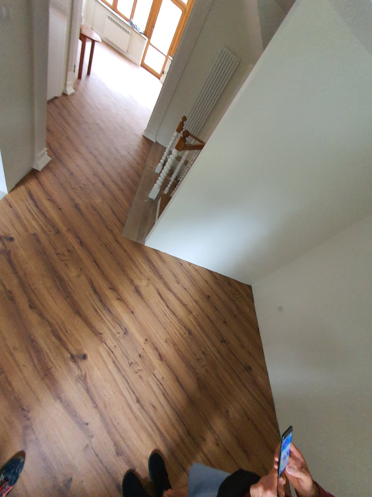 Nowo ułożone panele podłogowe w korytarzu, widok z góry z perspektywy osoby trzymającej telefon, schody z białymi balustradami w tle.