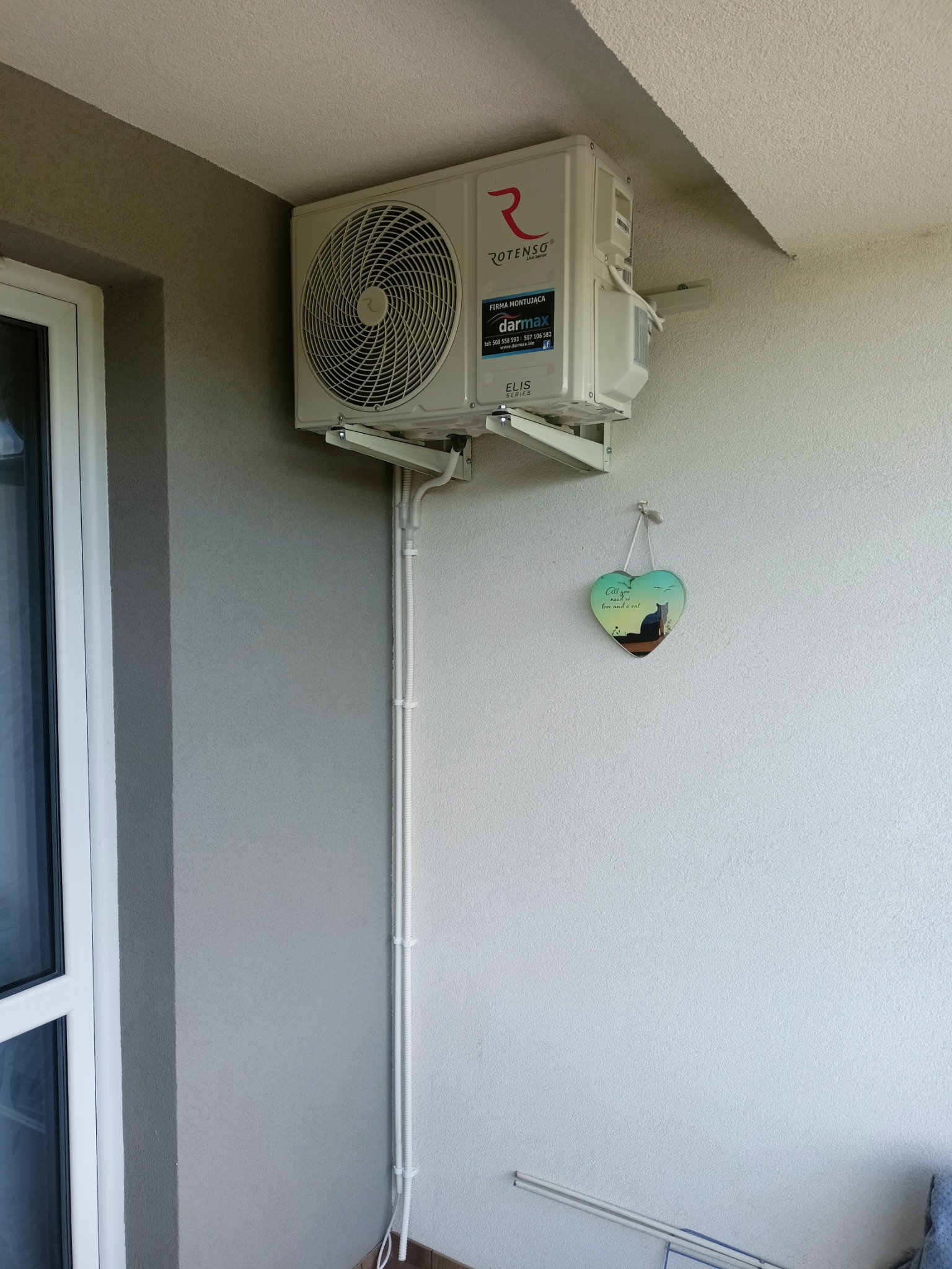 Zewnętrzna jednostka klimatyzacji Rotenso zamontowana na ścianie budynku, widoczne białe rurki instalacyjne i dekoracja ścienna w kształcie serca z motywem kota.