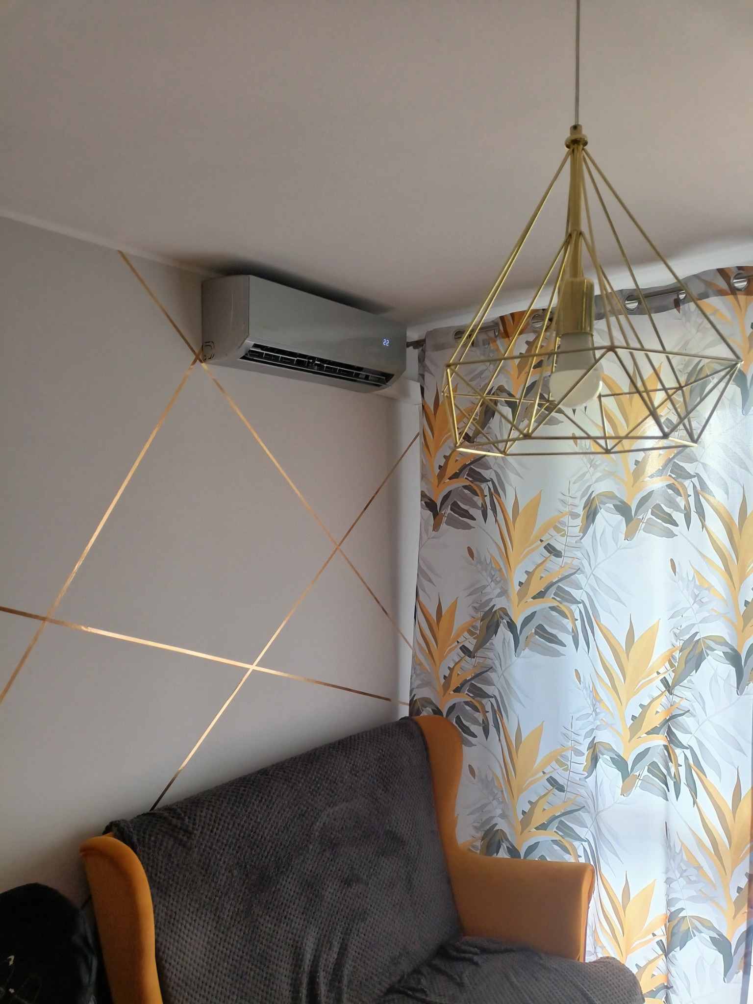 Szary klimatyzator ścienny zamontowany w pokoju z dekoracyjnymi złotymi paskami na ścianie, widoczna geometryczna lampa sufitowa i zasłony w roślinny wzór.