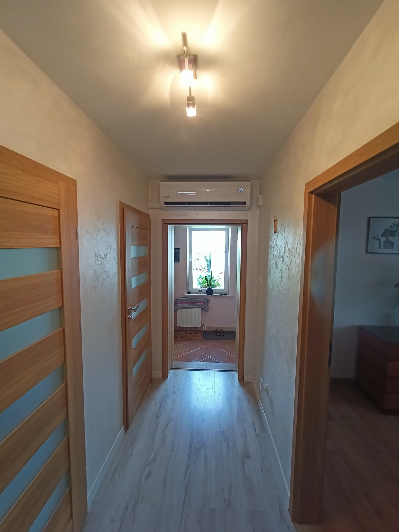 Klimatyzator marki KAISAI zamontowany nad drzwiami w korytarzu domu, widoczny wyświetlacz z temperaturą 23 stopnie Celsjusza, podłoga z paneli w odcieniach szarości.