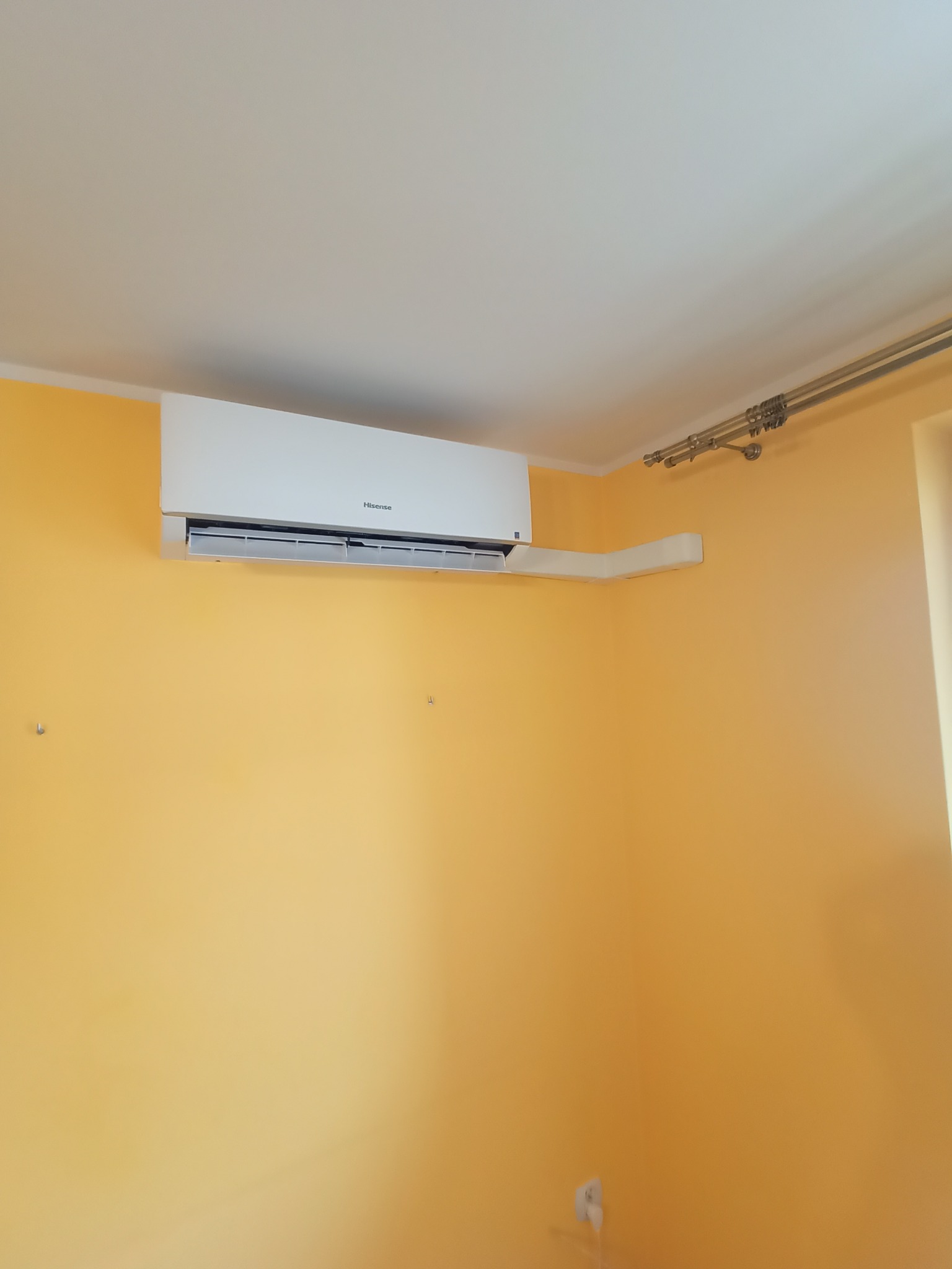 Biały klimatyzator Hisense zamontowany na pomarańczowej ścianie w pomieszczeniu z karniszem
