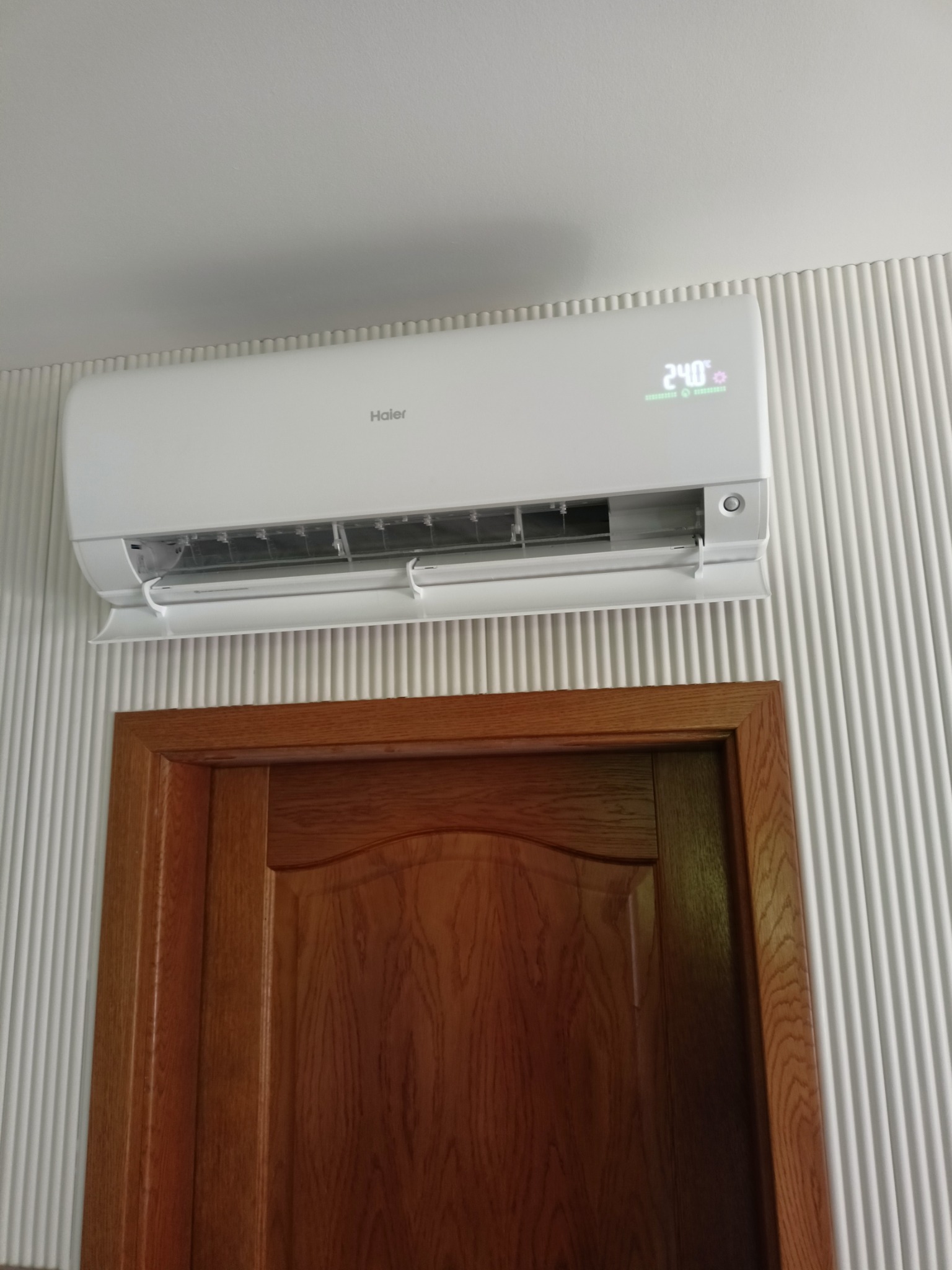 Biały klimatyzator Haier zamontowany nad drewnianymi drzwiami na ścianie z dekoracyjnymi pionowymi żłobieniami, wyświetlający temperaturę 24 stopnie Celsjusza.