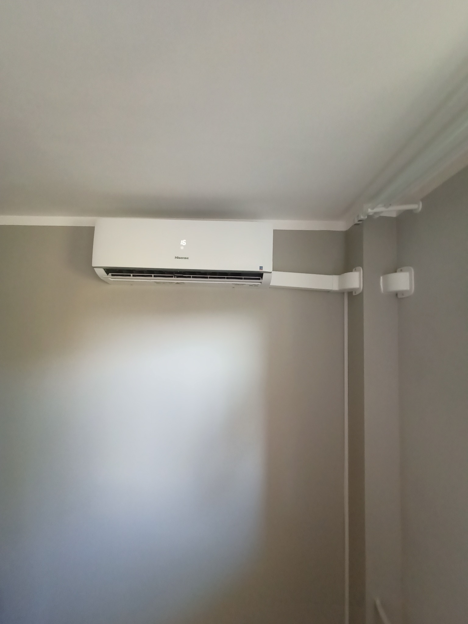 Biała jednostka klimatyzacji marki Hisense zamontowana na ścianie w pokoju o szarych ścianach, z widocznymi białymi kanałami i uchwytami na ścianie.