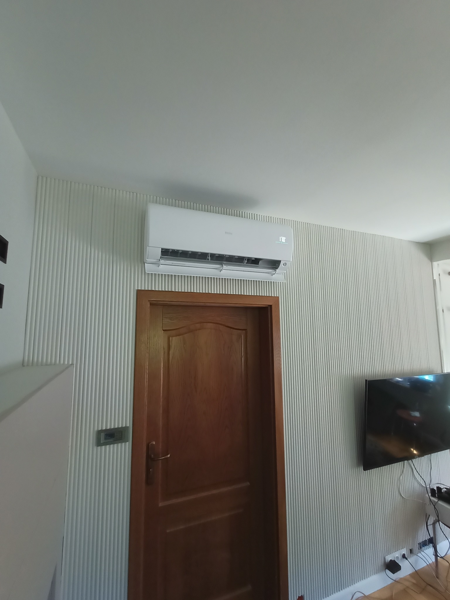 Biała klimatyzacja zamontowana nad drewnianymi drzwiami na ścianie z dekoracyjnymi pionowymi żłobieniami w jasnym pomieszczeniu.