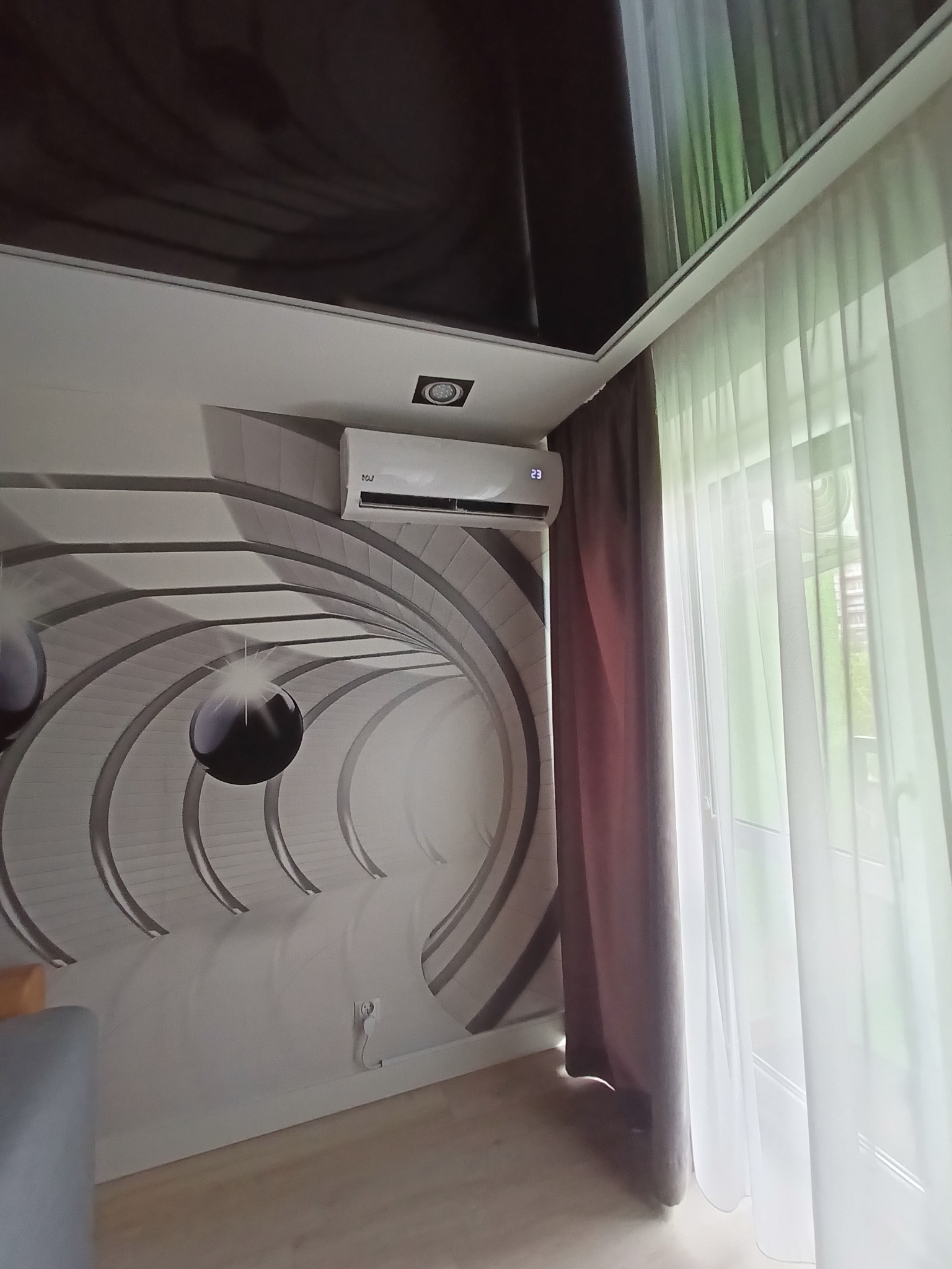 Biała klimatyzacja ścienna zamontowana w pokoju z tapetą z motywem tunelu i błyszczącym, ciemnym sufitem podwieszanym, obok okna z białymi zasłonami i brązowymi kotarami.