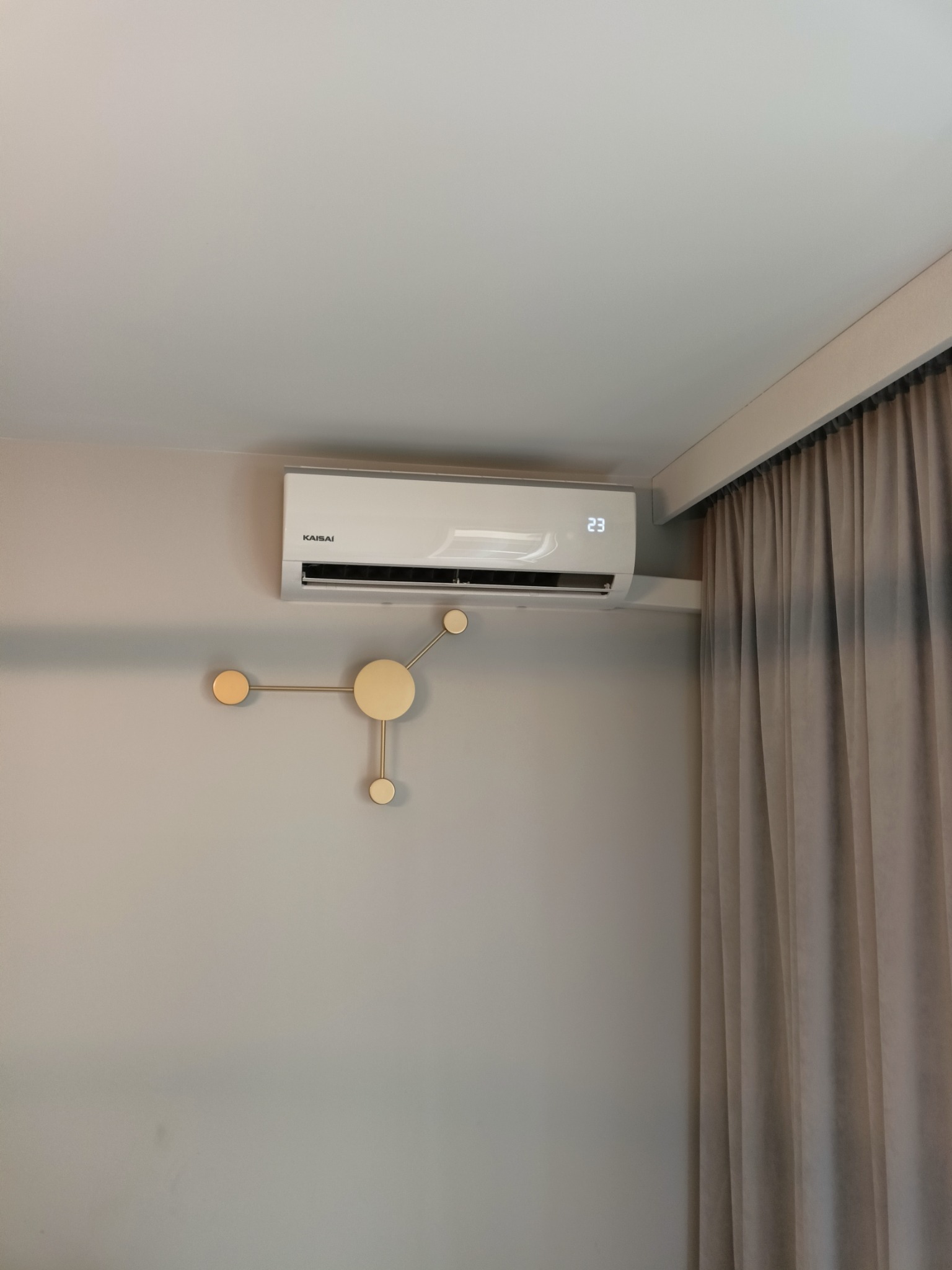 Biały klimatyzator Kaisai z wyświetlaną temperaturą 23 stopnie, zamontowany na szarej ścianie obok dekoracji ściennej w stylu molekularnym i zasłon w odcieniach beżu.