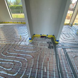 INSTALPRO Andrzej Kuczaj - Białe rury ogrzewania podłogowego ułożone na folii z czerwoną siatką w surowym wnętrzu z oknem dachowym, zabezpieczone taśmą żółtą i niebieską wzdłuż ścian.