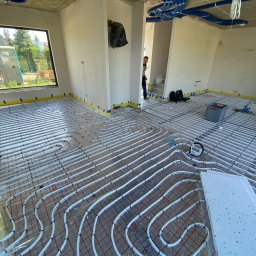 INSTALPRO Andrzej Kuczaj - Rozłożone rury ogrzewania podłogowego na folii z siatką, widoczne narzędzia i skrzynka, w tle okno z widokiem na przenośną toaletę na zewnątrz budynku w stanie surowym.