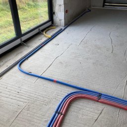 INSTALPRO Andrzej Kuczaj - Wrocław: Instalacja rur w podłodze w trakcie budowy, widoczne rury w kolorach niebieskim i czerwonym, prowadzone wzdłuż okna i ściany z pustaków.