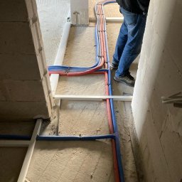 INSTALPRO Andrzej Kuczaj - Instalacja wodna i grzewcza w surowym stanie, rury PEX w kolorach niebieskim i czerwonym prowadzone po podłodze, mocowane do białej konstrukcji nośnej, widoczne fragmenty ścian z betonu komórkowego.