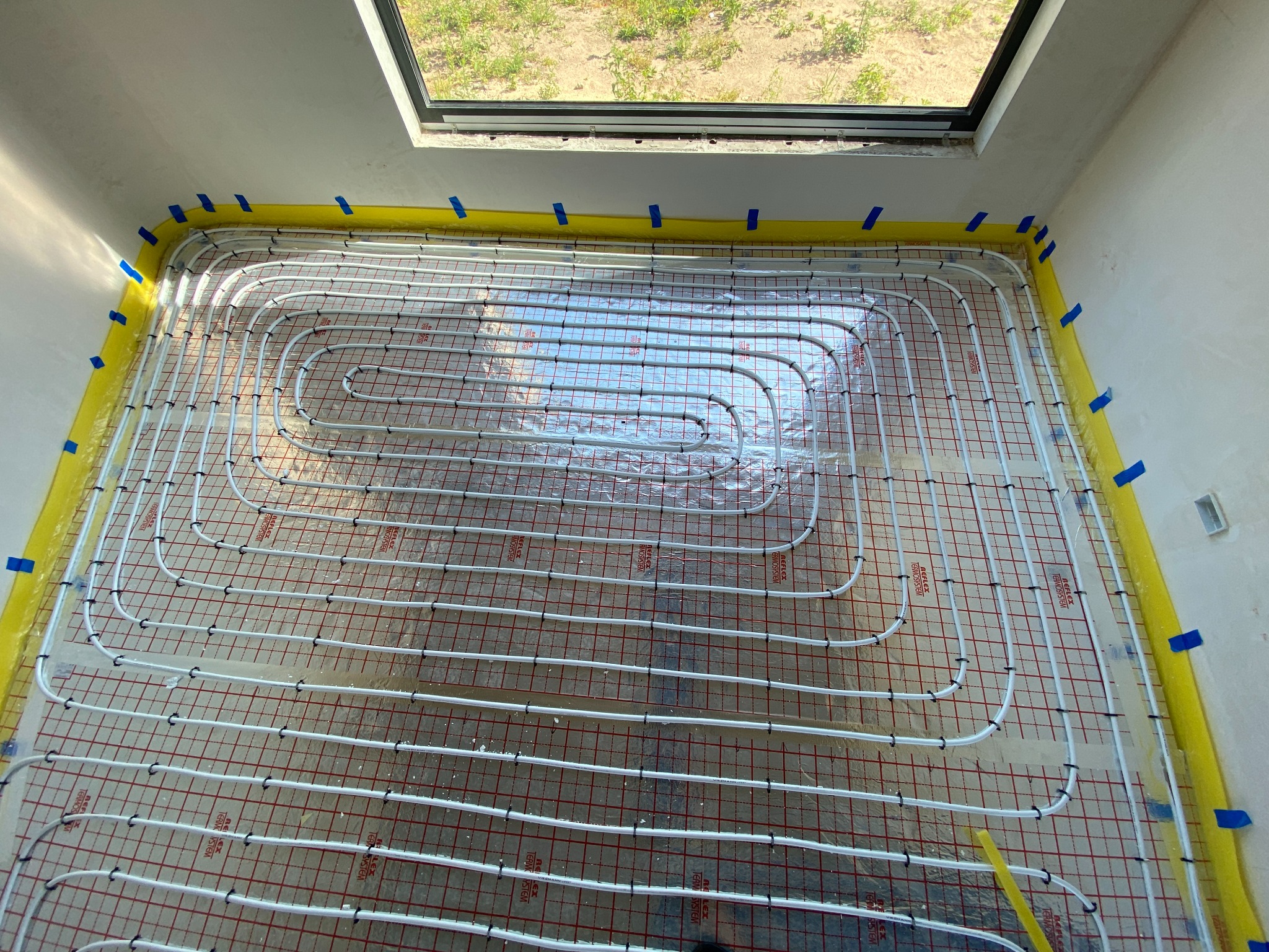 Białe rury ogrzewania podłogowego ułożone na folii z czerwoną kratką, zabezpieczone żółtą pianką i niebieską taśmą przy ścianach w surowym wnętrzu z oknem dachowym.