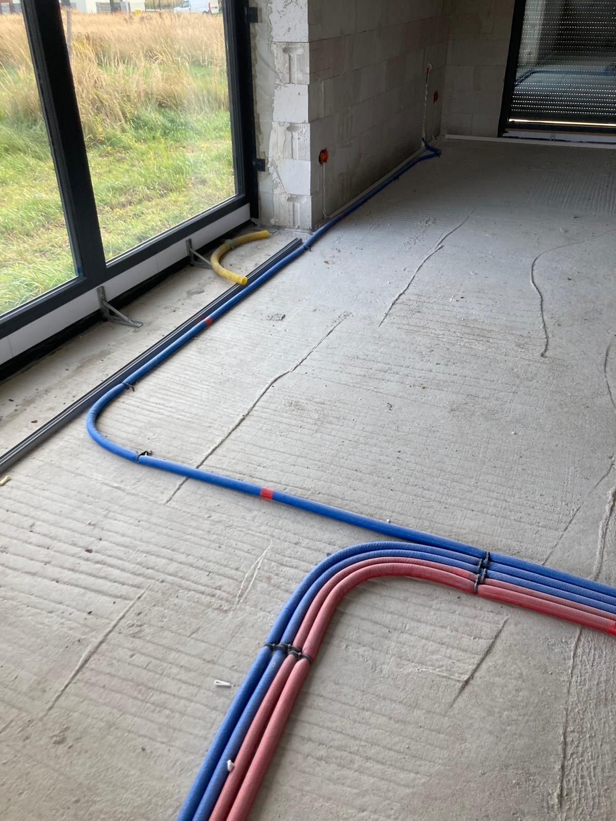 Wrocław: Instalacja rur w podłodze w trakcie budowy, widoczne rury w kolorach niebieskim i czerwonym, prowadzone wzdłuż okna i ściany z pustaków.