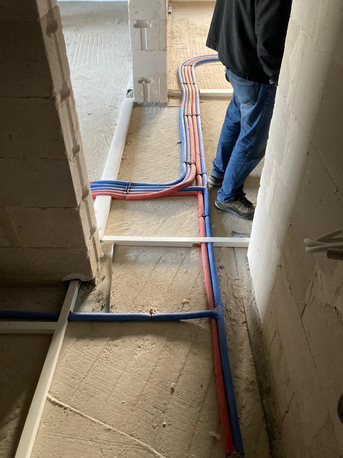 Instalacja wodna i grzewcza w surowym stanie, rury PEX w kolorach niebieskim i czerwonym prowadzone po podłodze, mocowane do białej konstrukcji nośnej, widoczne fragmenty ścian z betonu komórkowego.