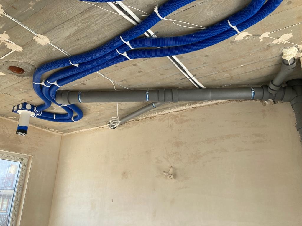 Niebieskie karbowane rury wentylacyjne podwieszone pod betonowym sufitem w surowym wnętrzu, szare rury odpływowe po prawej stronie, widoczne kable elektryczne i fragment okna.