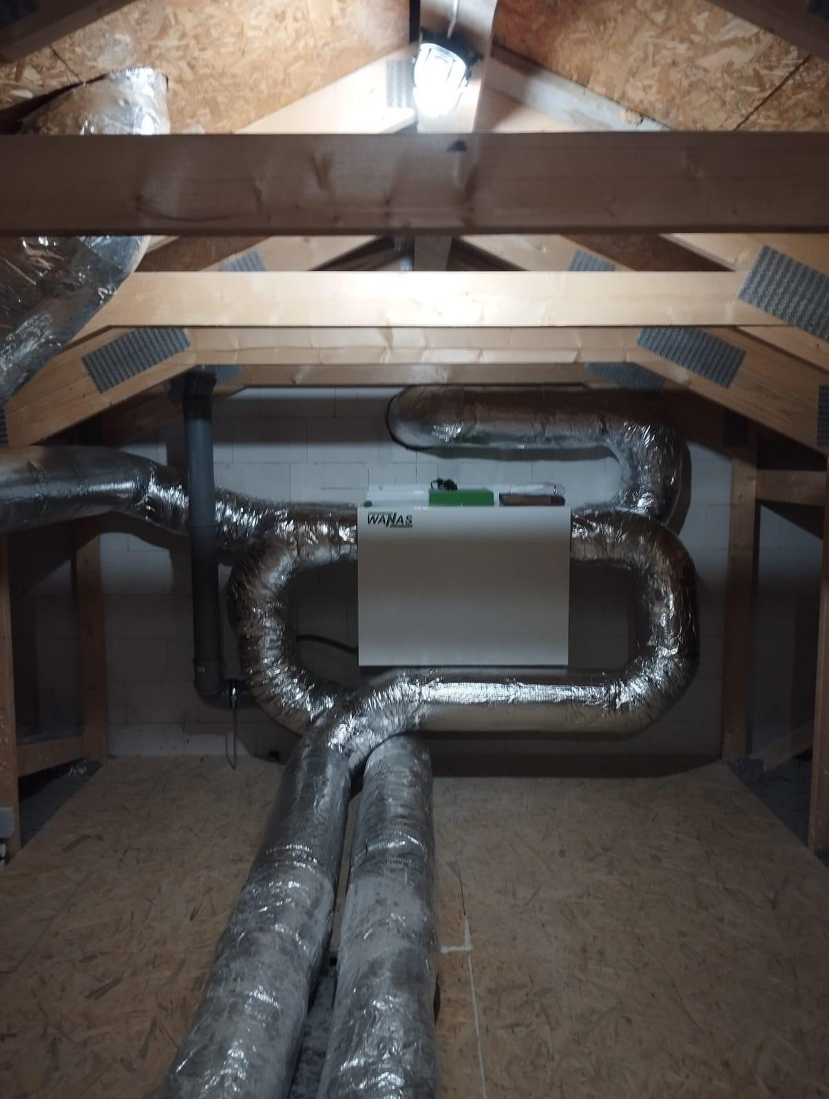 Centrala wentylacyjna WANAS z widocznymi przewodami wentylacyjnymi w izolacji aluminiowej, zainstalowana na poddaszu z drewnianą konstrukcją dachu i oświetleniem.