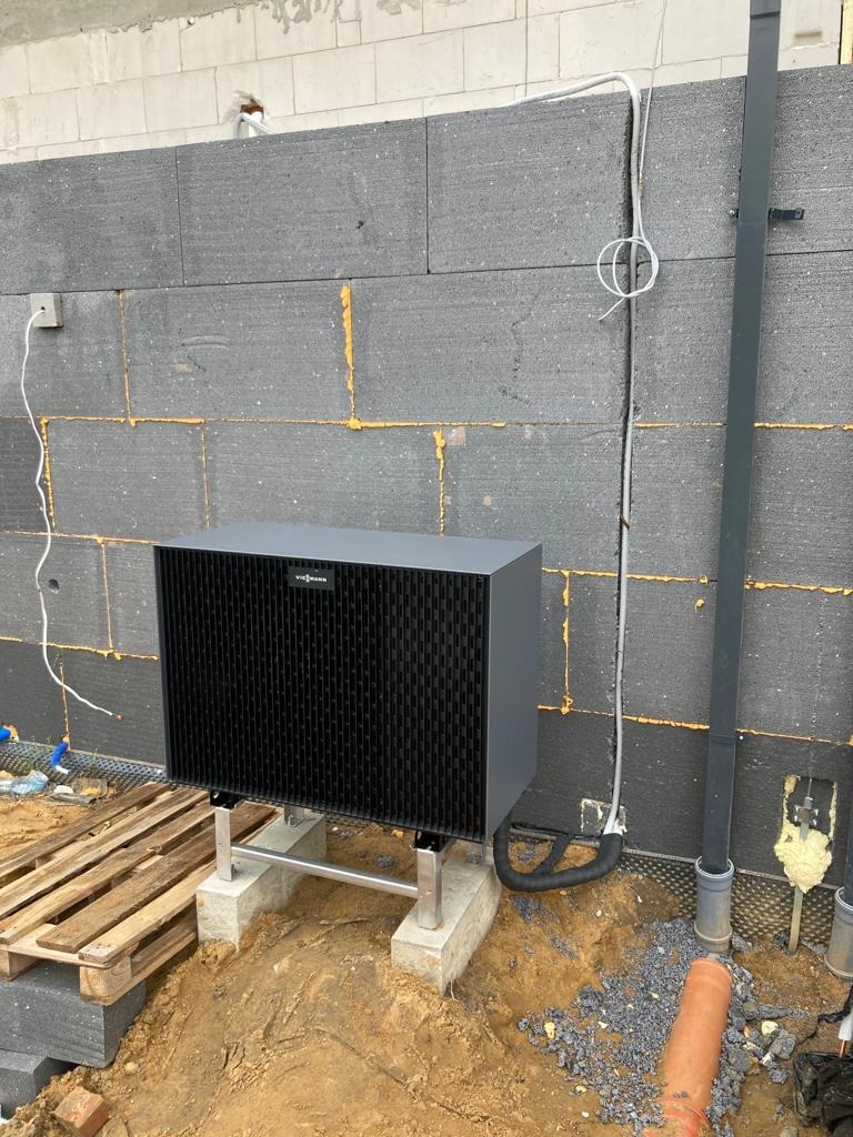 Czarna pompa ciepła Viessmann na betonowych podstawach, instalowana przy ścianie z izolacją styropianową, widoczne przewody i rury instalacyjne.