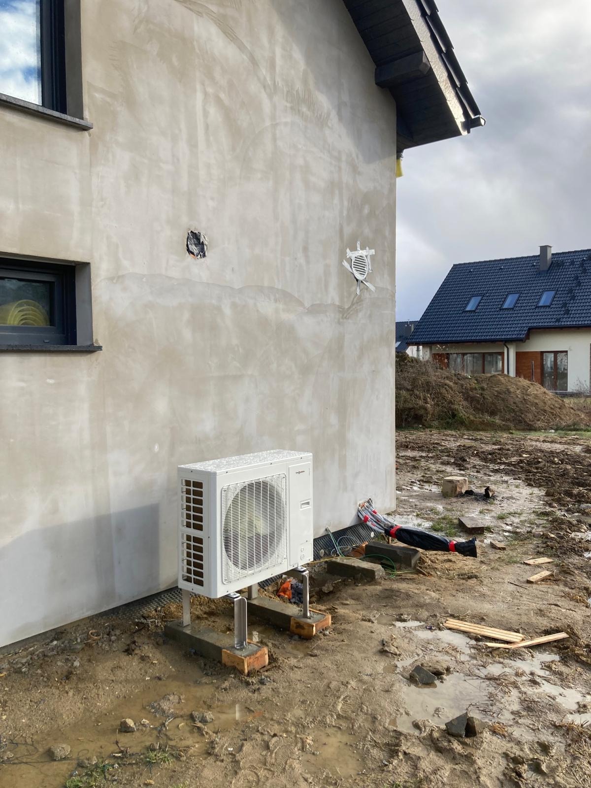 Biała jednostka zewnętrzna pompy ciepła Viessmann zamontowana na betonowych podstawach z cegłami, widoczne przewody w czarnej osłonie oraz otwory w surowej elewacji budynku w trakcie budowy,...