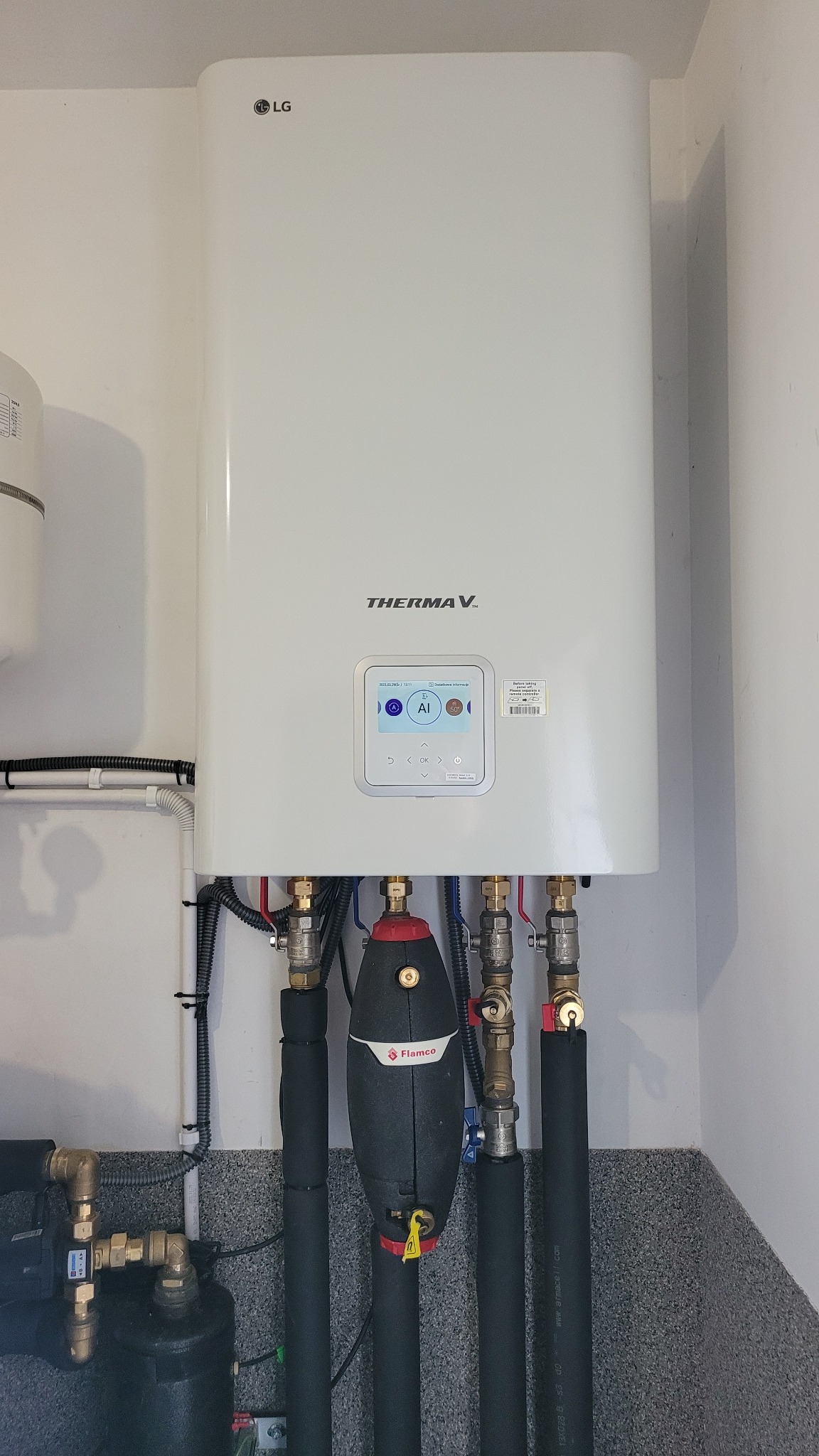 Biały piec gazowy LG Therma V z wyświetlaczem AI, podłączony do instalacji z rurami w izolacji, widoczny także zawór Flamco.