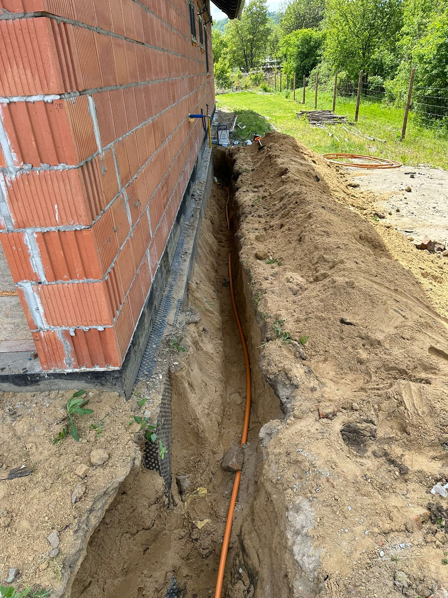 Pomarańczowa rura gazowa ułożona w wykopie wzdłuż ściany budynku z cegły ceramicznej, widoczne przyłącze z zaworem, teren budowy z ziemią i roślinnością.