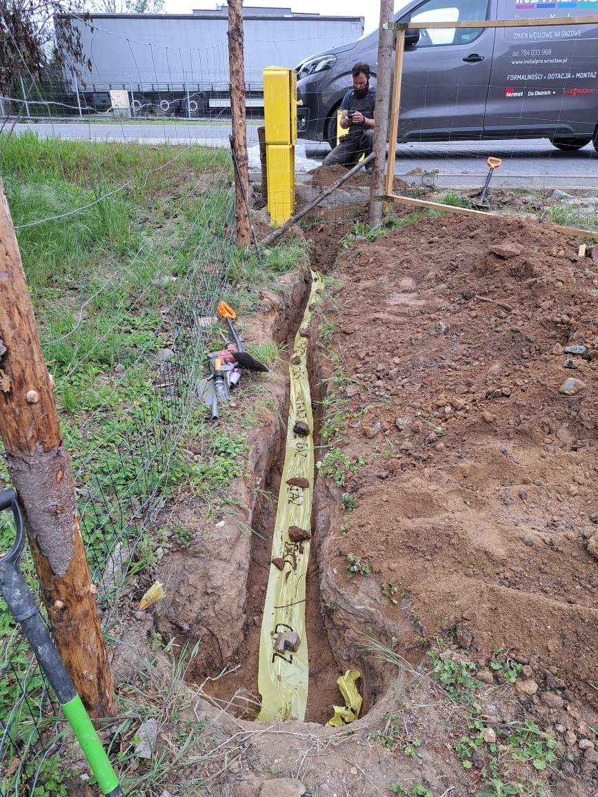 Wykop pod rurę gazową zabezpieczony żółtą folią ochronną z napisem GAZ, widoczne narzędzia pracy, pracownik siedzący obok samochodu firmy zajmującej się instalacjami gazowymi.