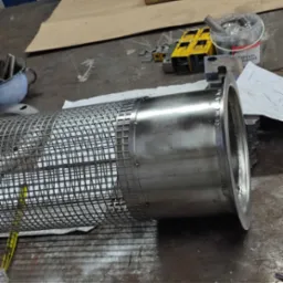 Metalowy cylinder z siatką ochronną, spawany pierścień mocujący, widoczne ślady obróbki na stole warsztatowym, narzędzia i akcesoria w tle.