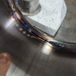 Ślusarskie usługi mobilne Łukasz Piekielniak - Detal spawanego, nierdzewnego pierścienia z widocznymi śladami spawania i przebarwieniami termicznymi, obok metalowy cylinder i porowaty blok