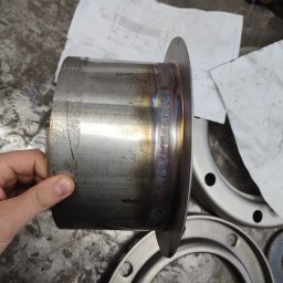 Ślusarskie usługi mobilne Łukasz Piekielniak - Metalowy kołnierz z widocznymi śladami spawania, trzymany w dłoni, na tle warsztatowego stołu z planami technicznymi i innymi elementami metalowymi.