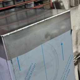 Ślusarskie usługi mobilne Łukasz Piekielniak - Szczegółowo sfotografowane, spawane połączenie blachy nierdzewnej z widocznymi śladami spawania, zabezpieczone folią ochronną z nadrukiem w tle warsztat z półkami na narzędzia.