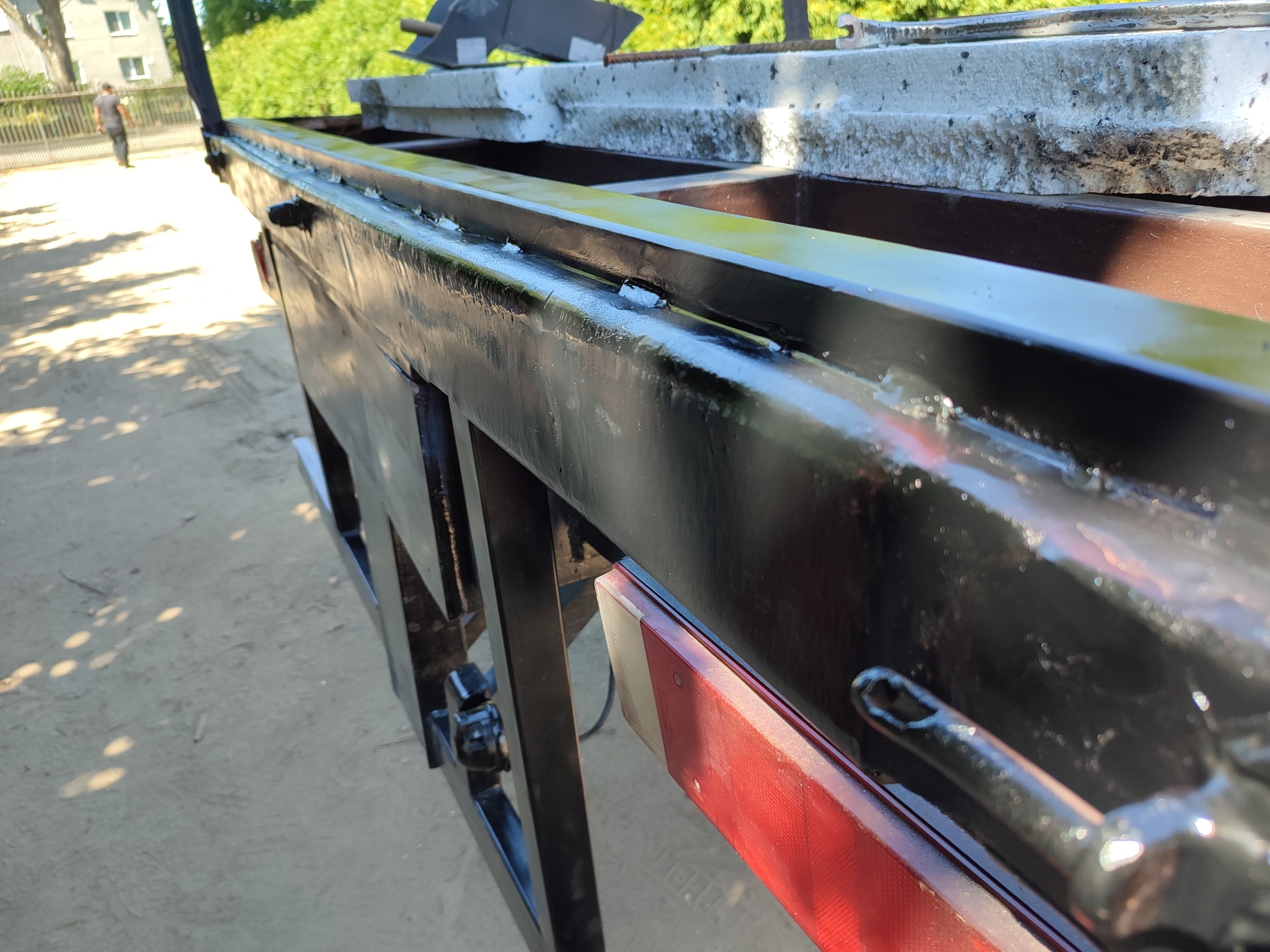 Fragment konstrukcji metalowej przyczepy z widocznymi spawami, świeżo pomalowana na czarno, z elementami odblaskowymi i betonowymi płytami na górze, sfotografowana z bliska.