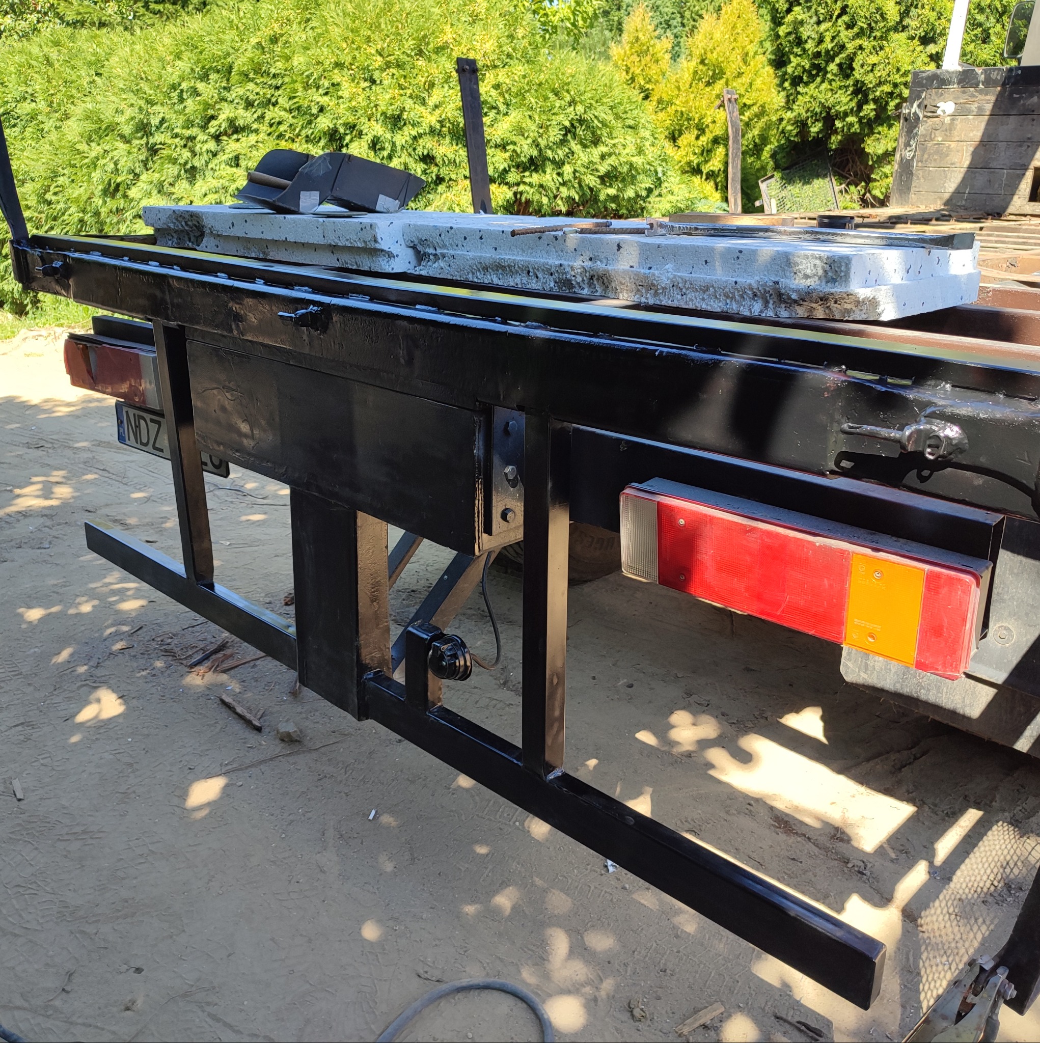 Tylny widok nowo pomalowanej, czarnej metalowej konstrukcji przyczepy z zamontowanymi światłami, przewożącej betonowe elementy, na tle zieleni.