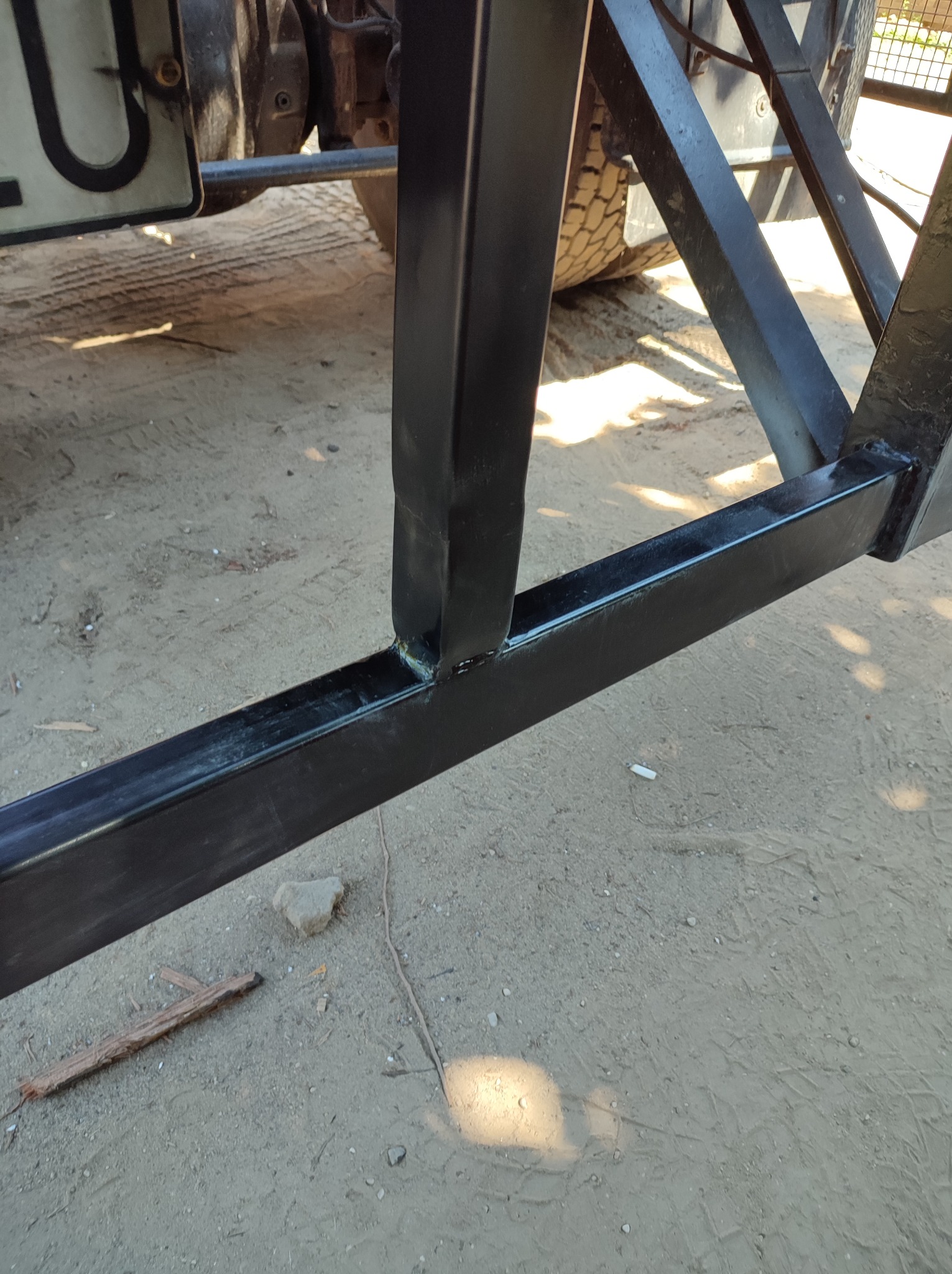 Fragment spawanej, czarnej konstrukcji metalowej przyczepy, widoczne spawy, opona i tablica rejestracyjna w tle, na piaszczystym podłożu.