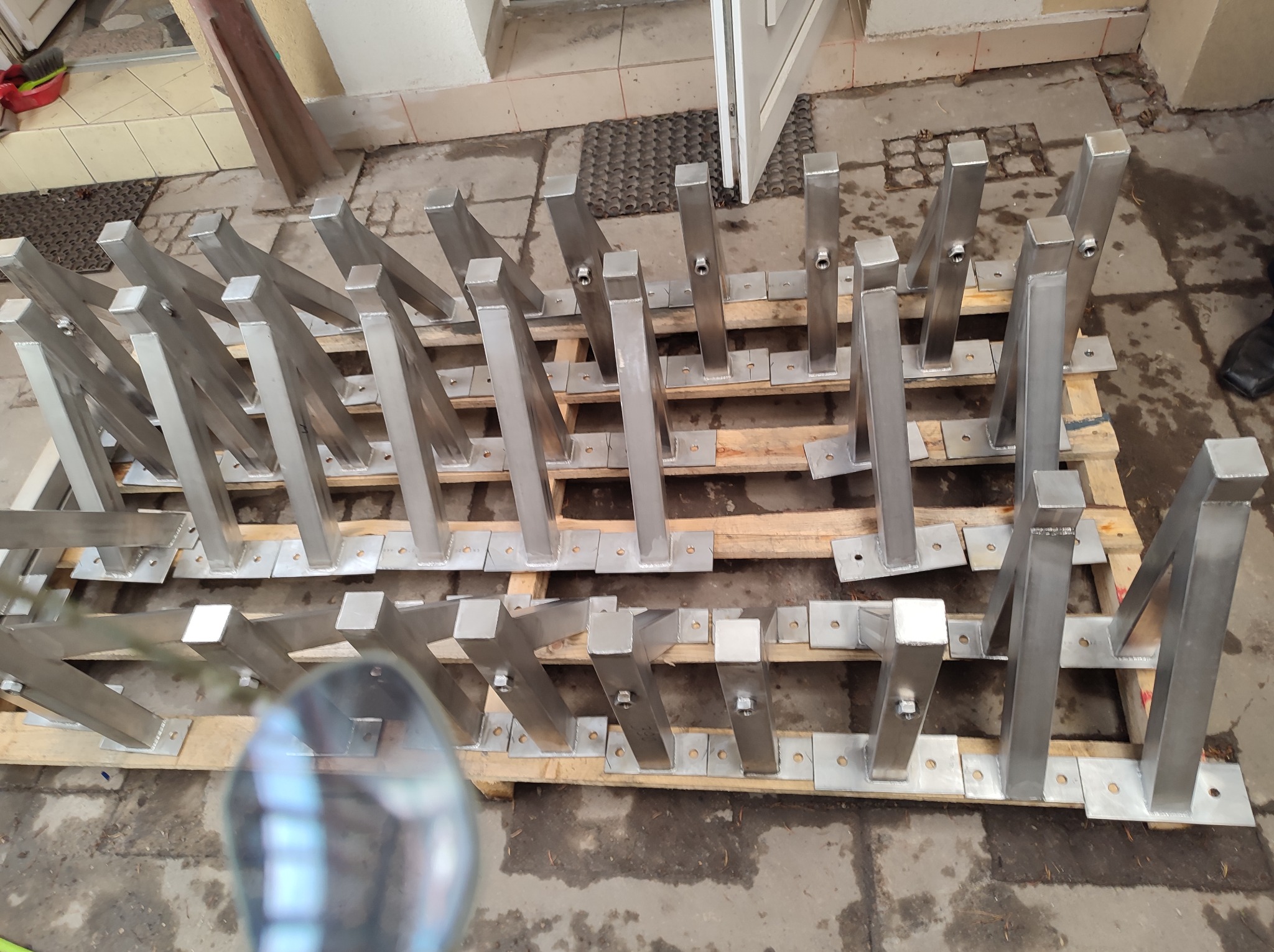 Metalowe wsporniki balustrad lub poręczy, ułożone na drewnianej palecie, widoczne ślady spawania i otwory montażowe.