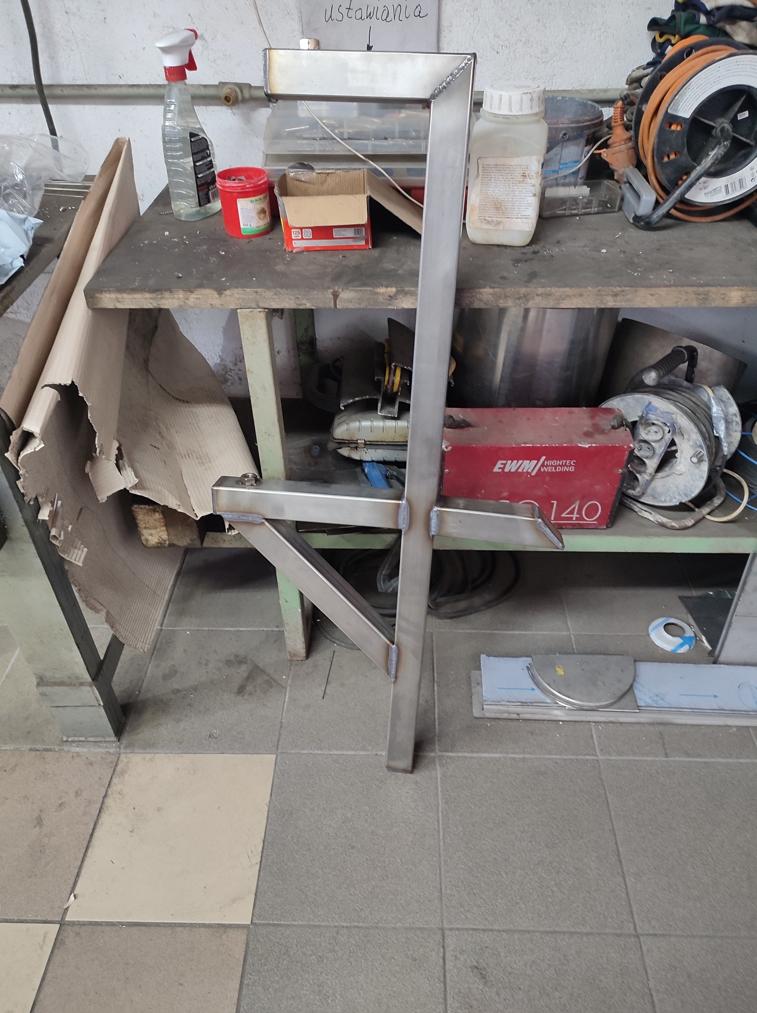 Metalowa konstrukcja wspornikowa z profili spawanych, stojąca na podłodze warsztatu, widoczna spawarka EWM i narzędzia na stole roboczym w tle.