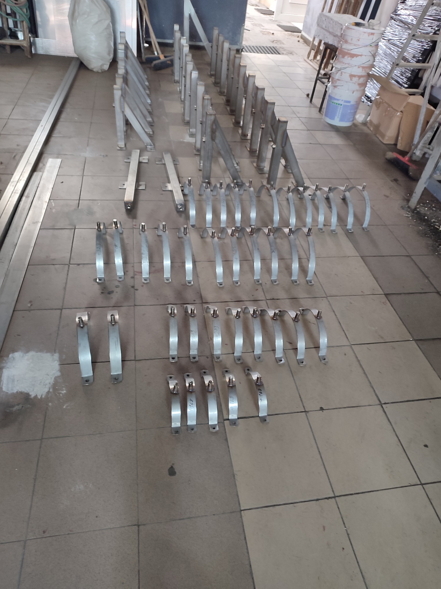 Przygotowane do montażu metalowe elementy konstrukcyjne, prawdopodobnie do balustrad lub poręczy, ułożone rzędami na podłodze warsztatu, widoczne ślady obróbki, niektóre z naniesionymi numerami