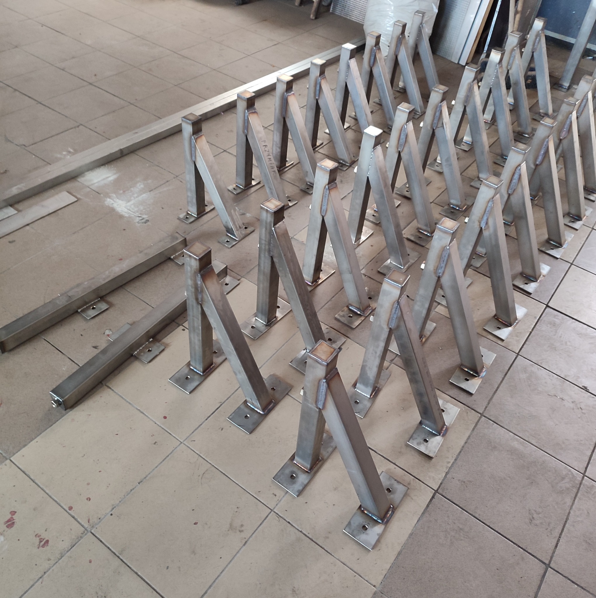 Metalowe podpory balustrad lub poręczy, spawane, z podstawami do mocowania, ułożone na podłodze warsztatu przed montażem.