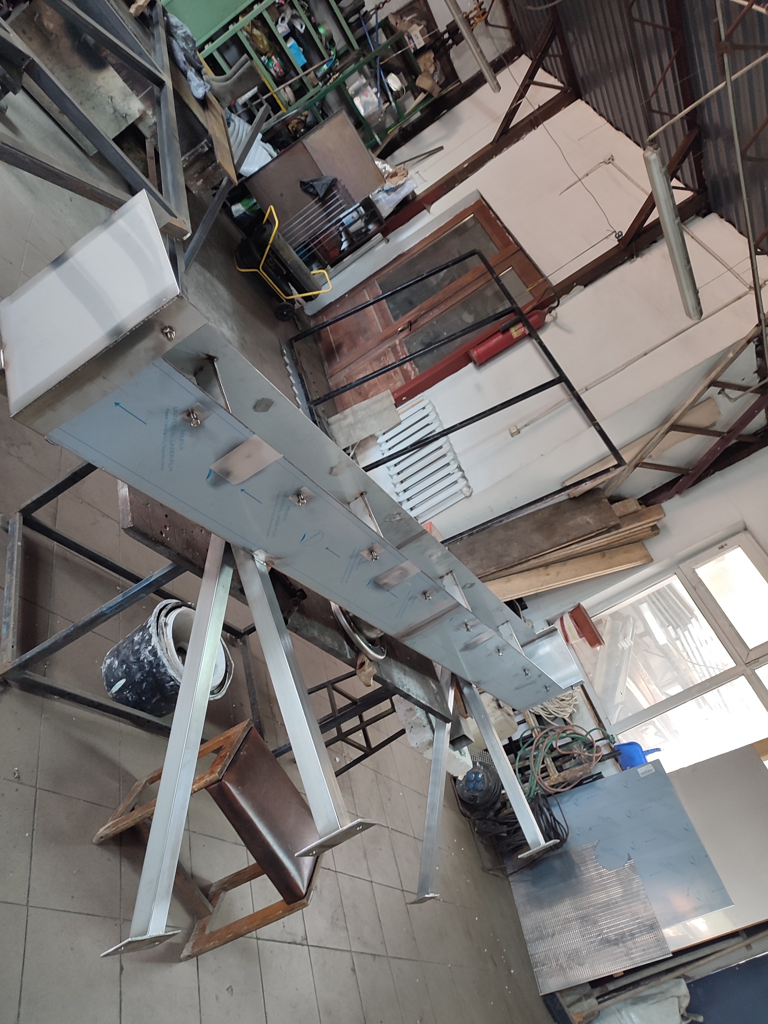 Wnętrze warsztatu z elementem konstrukcyjnym z blachy nierdzewnej na metalowym stelażu, widoczne ślady obróbki i montażu, tło warsztatowe z narzędziami i materiałami.