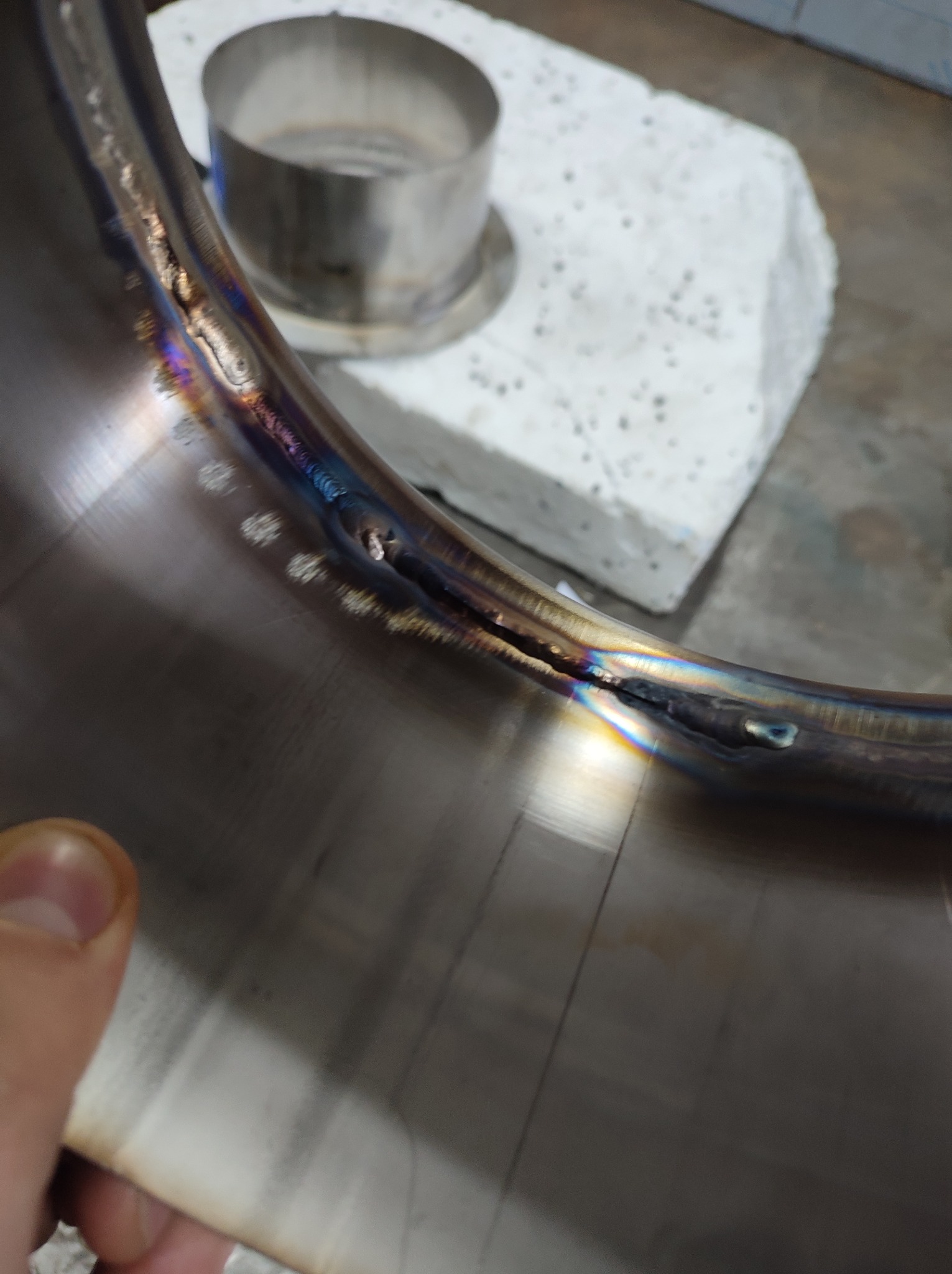 Detal spawanego, nierdzewnego pierścienia z widocznymi śladami spawania i przebarwieniami termicznymi, obok metalowy cylinder i porowaty blok