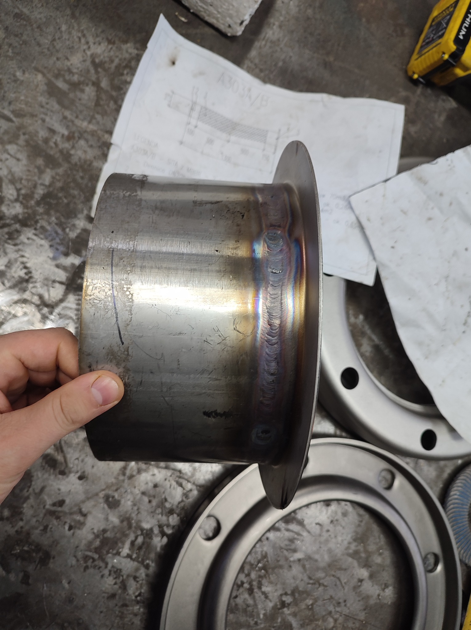 Metalowy kołnierz z widocznymi śladami spawania, trzymany w dłoni, na tle warsztatowego stołu z planami technicznymi i innymi elementami metalowymi.