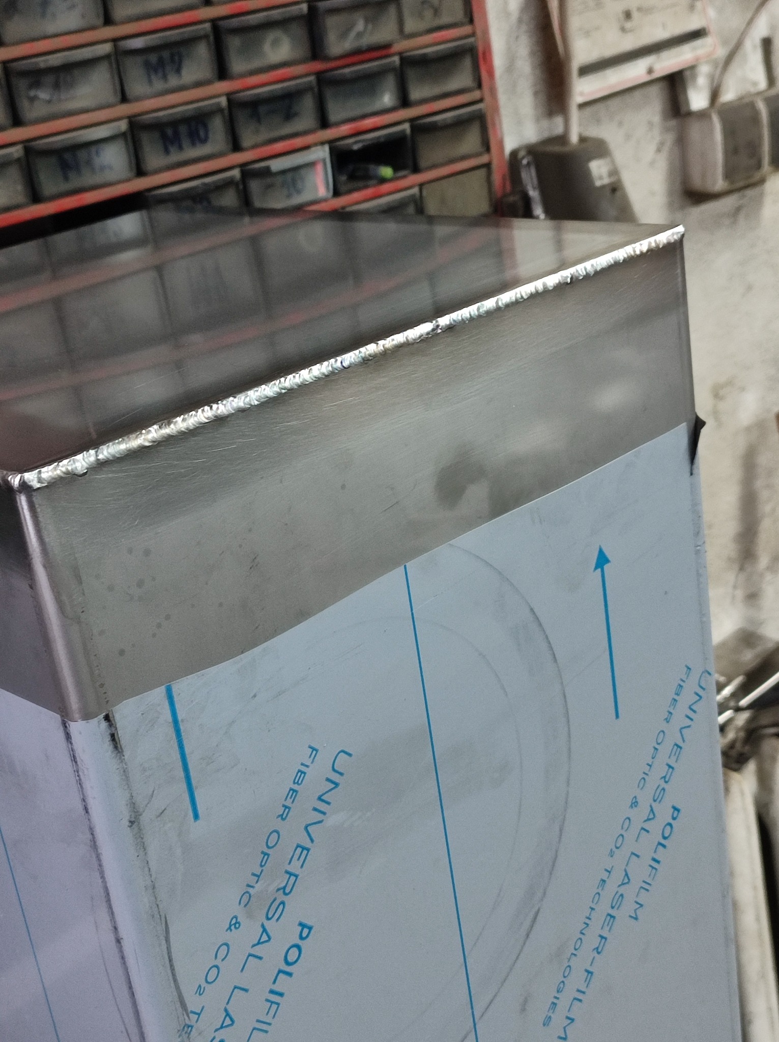 Szczegółowo sfotografowane, spawane połączenie blachy nierdzewnej z widocznymi śladami spawania, zabezpieczone folią ochronną z nadrukiem w tle warsztat z półkami na narzędzia.