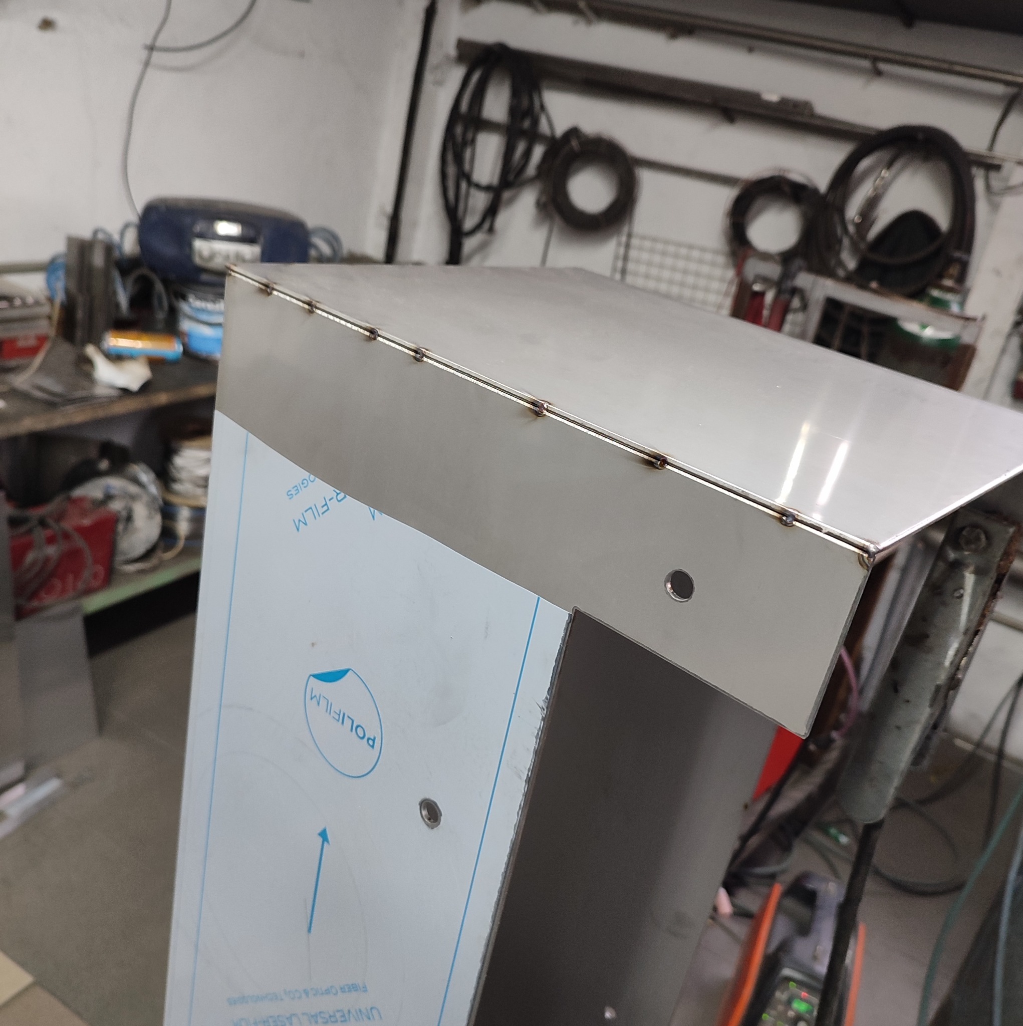 Detal metalowej konstrukcji z widocznymi spawami i otworami, pokrytej folią ochronną, w warsztatowym otoczeniu.