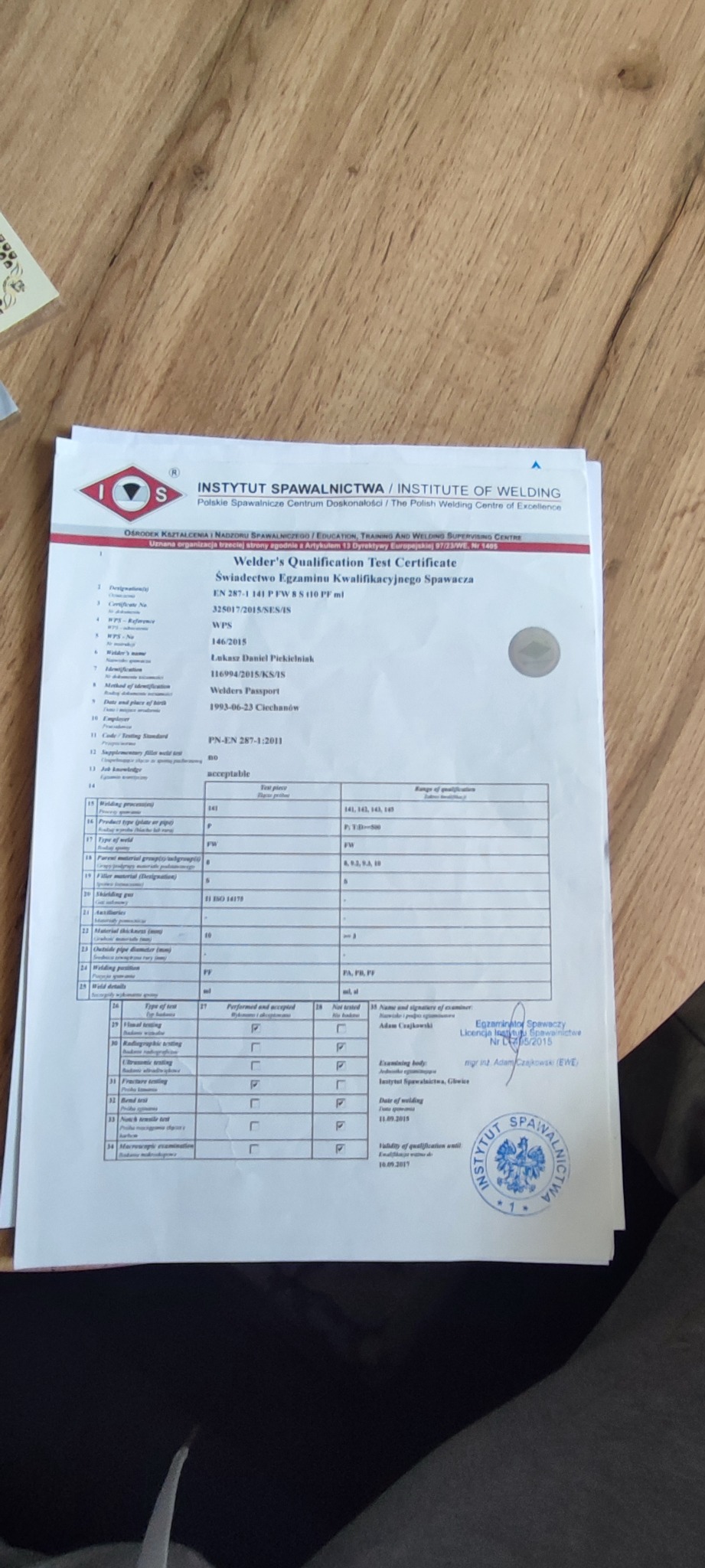 Skan certyfikatu kwalifikacyjnego spawacza z Instytutu Spawalnictwa, widoczny stempel instytucji i podpisy egzaminatorów, leżący na drewnianej powierzchni.