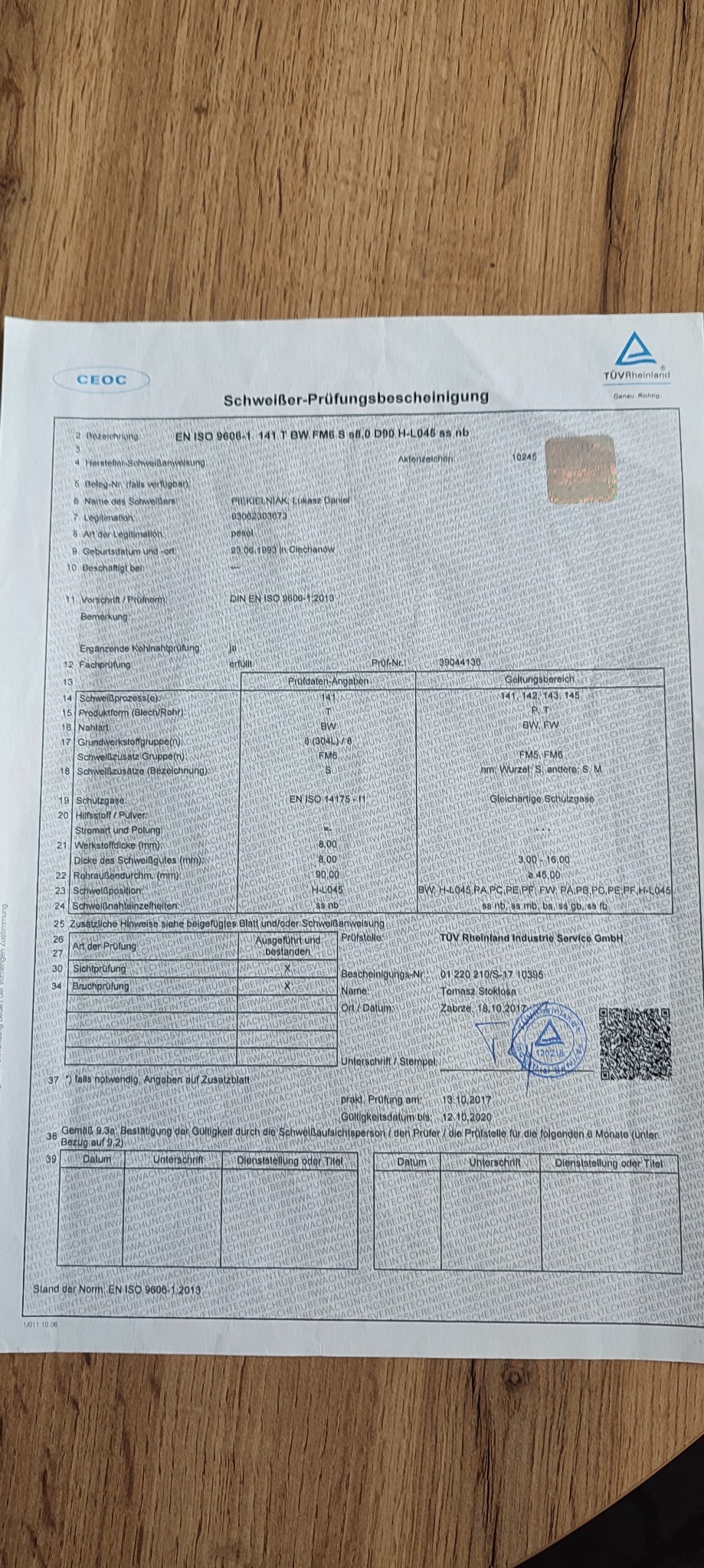 Skan certyfikatu spawacza, wydanego przez TÜV Rheinland, z danymi osobowymi i zakresem uprawnień, leżący na drewnianej powierzchni.