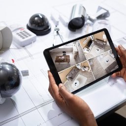 Elektrokol - Na stole z planami leżą kamery monitoringu, centrala alarmowa i tablet wyświetlający podgląd z czterech kamer w różnych pomieszczeniach.