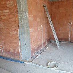 Elektrokol - Wnętrze w trakcie prac wykończeniowych: ściany z cegły dziurawki, surowy betonowy filar, drabina oparta o ścianę, widoczne przewody elektryczne wychodzące z puszek instalacyjnych, niebieska rura...