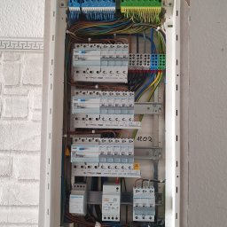 Elektrokol - Otwarta skrzynka z zabezpieczeniami elektrycznymi Hager, widoczne różnokolorowe przewody, bezpieczniki i listwy zaciskowe na tle fragmentu otynkowanej ściany.